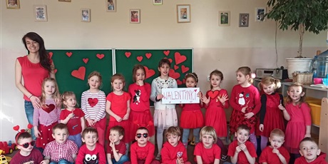 Powiększ grafikę: Nauczycielka z grupą dzieci pozuje do zdjęcie. Są ubrani na czerwono. Dwie dziewczynki trzymają kartę z napisem "Walentynki"