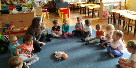 Powiększ grafikę: Dzieci z nauczycielką siedzą na dywanie. Po środku koła leży lalka i samochodzik