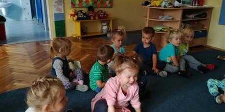 Powiększ grafikę: Dzieci siedzą na dywanie i patrzą się na siebie wzajemnie. Dziewczynka w różowej bluzce próbuje wstać