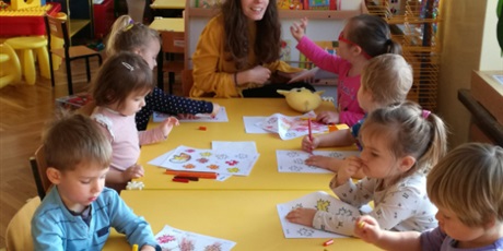 Powiększ grafikę: Dzieci siedzą z nauczycielką przy stolikach. Kolorują karty pracy
