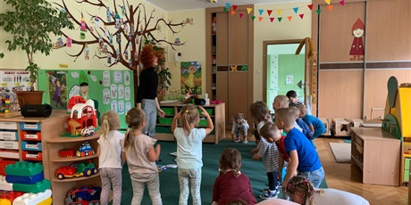 Powiększ grafikę: Dzieci stoją na końcu dywanu. Po środku stoi nauczycielka i skacze
