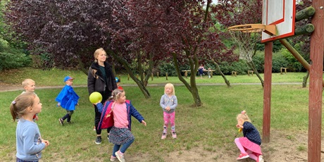 Powiększ grafikę: Dziewczynka próbuje rzucić piłką do kosza. Wokół niej stoją dzieci. Za dziewczynką stoi dorosła kobieta