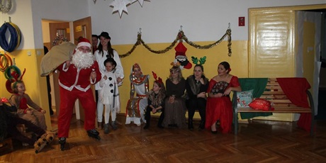 Powiększ grafikę: Mikołaj wchodzi do sali. Z boku siedzą przebrani dorośli