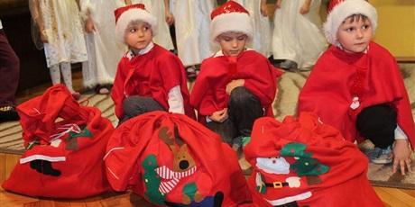 Powiększ grafikę: Trzech chłopców kuca na podłodze. Mają na sobie czerwone peleryny i czapki Świętego Mikołaja. Przed nimi stoją czerwone worki