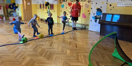 Powiększ grafikę: Czwórka dzieci stoi przed linią. Kopią piłki do bramki. Trener stoi obok dzieci