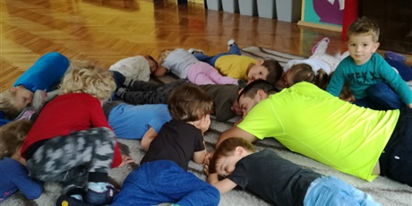 Powiększ grafikę: Dzieci wraz z trenerem leżą na dywanie. Chłopiec patrzy się i uśmiecha