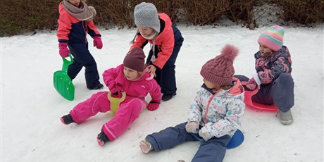 Powiększ grafikę: 3 dziewczynki w zimowych ubraniach przygotowuje się do zjady na plastikowych sankach. W tle dwie dziewczynki w różowo-granatowych kombinezonach