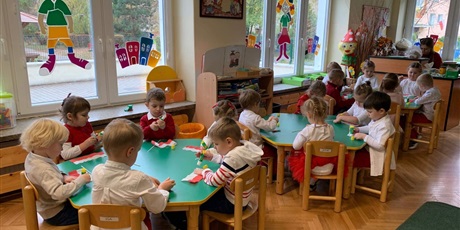 Powiększ grafikę: Dzieci siedzą przy stołach w sali przedszkolnej. Wykonują flagi Polski