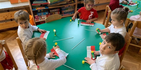 Powiększ grafikę: Piątka dzieci siedzi przy stole. Wykonują flagi Polski