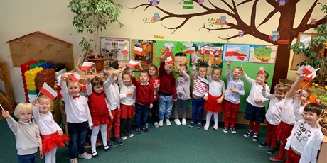 Powiększ grafikę: Dzieci stoją w półkolu. Machają zrobionymi przez siebie flagami Polski