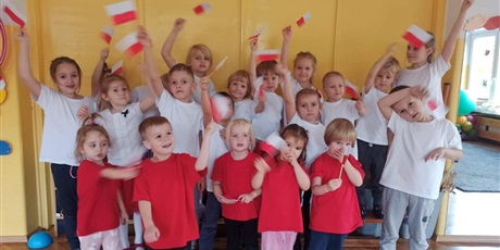 Powiększ grafikę: Grupa dzieci macha flagami Polski. Starsze dzieci mają białe koszulki. Młodsze dzieci mają czerwone koszulki