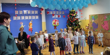 Powiększ grafikę: Nauczycielka wraz z dziećmi i rytmiczką stoją przed tłem z dekoracją
