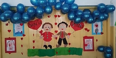 Powiększ grafikę: Ozdobiona ściana przedszkolna, na której znajdują się dwie postacie trzymające balony z napisem: babcia i dziadek. Na górze przyczepione są niebieskie balony