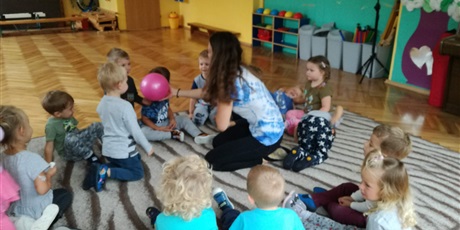 Powiększ grafikę: Dzieci siedzą na dywanie. Nauczycielka podaje dziecku różową piłkę