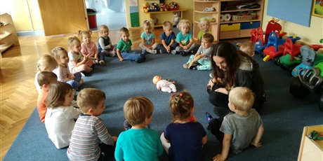 Powiększ grafikę: Dzieci z nauczycielką siedzą w kole na dywanie. Na dywanie po środku leży lalka