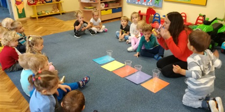 Powiększ grafikę: Dzieci siedza na dywanie z nauczycielką. Na dywanie położone są kolorowe kartki z przezroczystymi kubkami