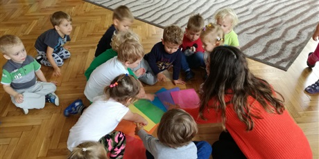 Powiększ grafikę: Dzieci z nauczycielką siedzą na sali gimnastycznej. Rozkładają na podłodze kolorowe kartki
