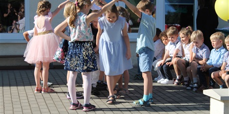Powiększ grafikę: Dzieci w parach podnoszą ręce do góry. Dziewczynka w niebieskiej sukience przechodzi pod utworzonym mostkiem. Na drugim planie młodsze dzieci oglądają występ