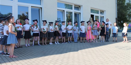 Powiększ grafikę: Grupa dzieci z czapki na głowie trzyma w rękach kwiaty. Dzieci śpiewają. W tle widać nauczycielki przedszkola