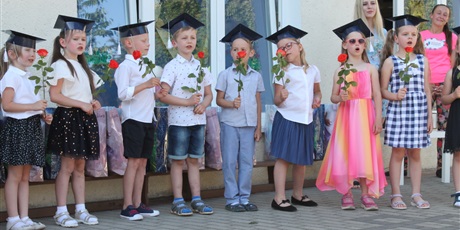 Powiększ grafikę: Osiem dzieci z czapkami na głowach trzyma w rękach kwiatek. Dzieci śpiewają. W tle widać dwie kobiety