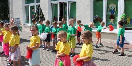 Powiększ grafikę: Dziewczynki w żółtych koszulkach trzymają pompony. Za nimi stoją chłopcy w zielonych koszulkach, trzymając w rękach pompony