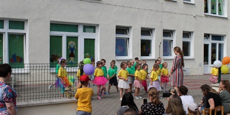 Powiększ grafikę: Dzieci w żółtych i zielonych koszulkach ustawiają się do przedstawienia. Przed nimi stoi dorosła kobieta. Przed nimi siedzą dorosłe osoby czekające na występ