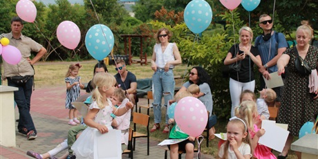 Powiększ grafikę: Dzieci trzymają balony niebieskie i różowe w kropki. W tle widać stojących, dorosłych ludzi